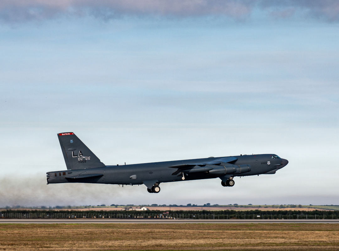 U.S. Air Force B-52 Stratotanker from Barksdale Air Force Base flying low above a runway with landing gear deployed, dark gray fuselage visible against a rural landscape and cloudy sky.