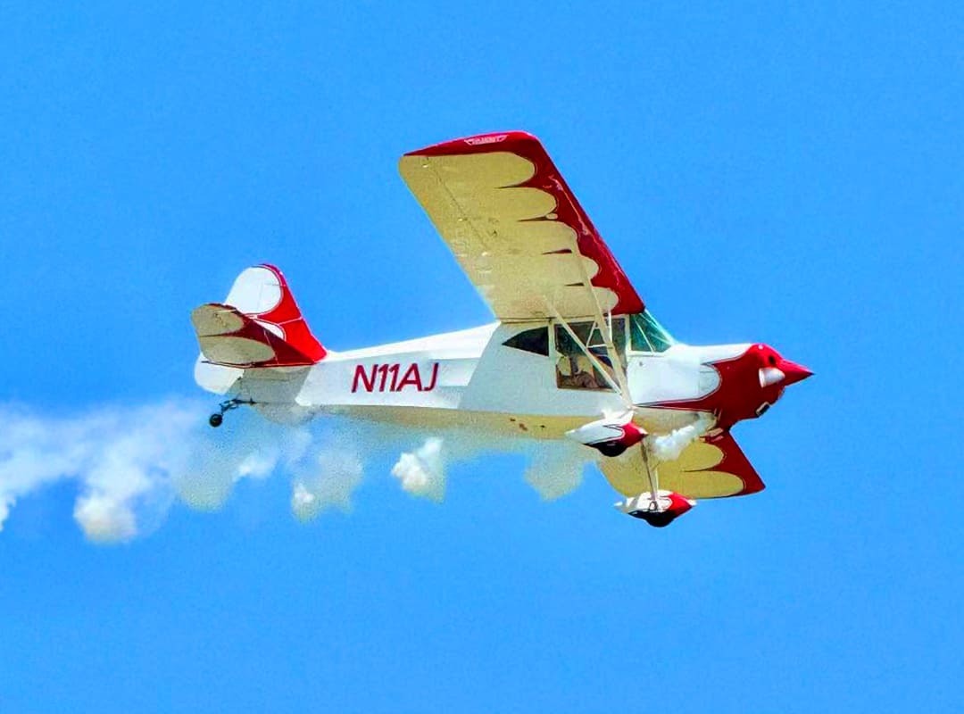Eric Hollingsworth flying a red and white Taylorcraft L-2 aircraft, banking in flight against a bright blue sky with a trail of white smoke behind it.
