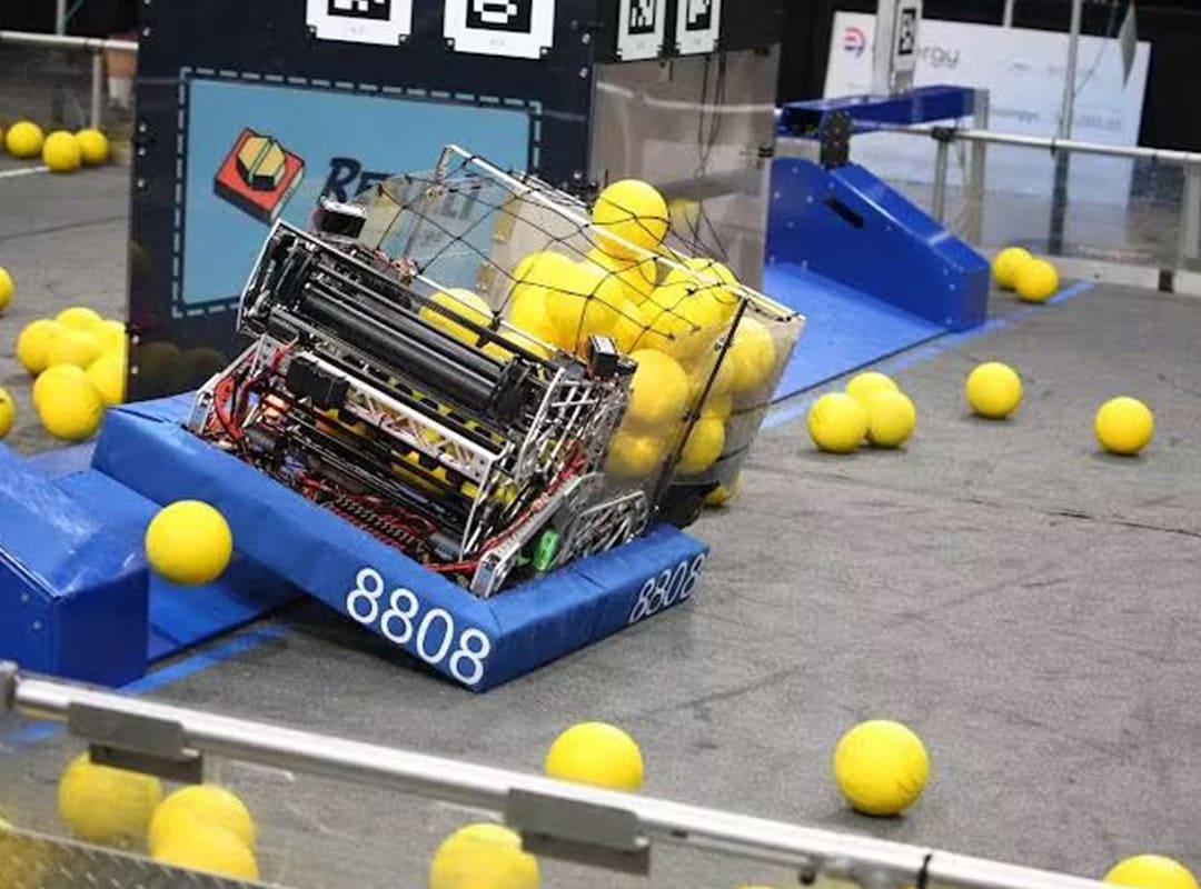 Student-built competition robot collects bright yellow balls on a robotics playing field during a STEM activity.