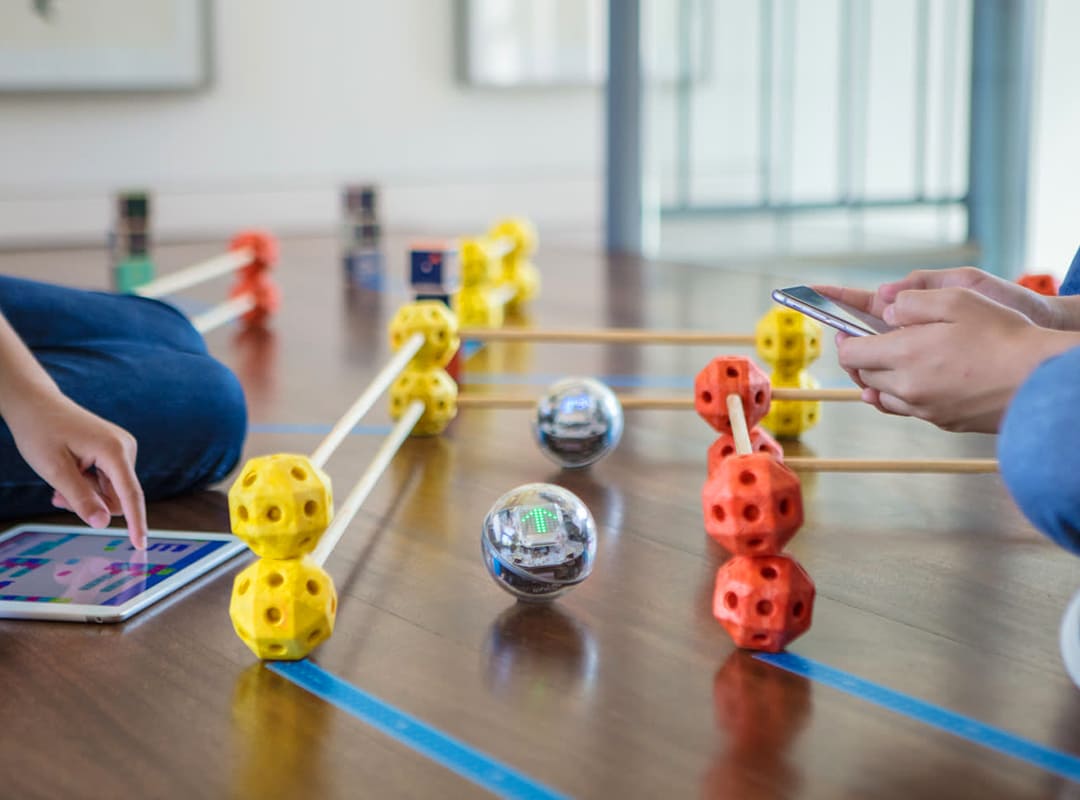 Students use tablets and smartphones to program and control small educational robots navigating a colorful obstacle course.