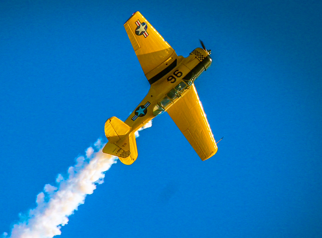Yellow T-6 Texan performing a steep bank against a bright blue sky, trailing white smoke during an aerobatic maneuver.