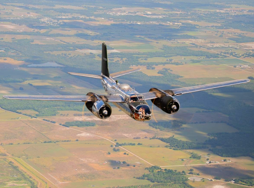 A twin-engine A-26 Invader flies over patchwork farmland, viewed from the front with its propellers spinning and nose art visible.